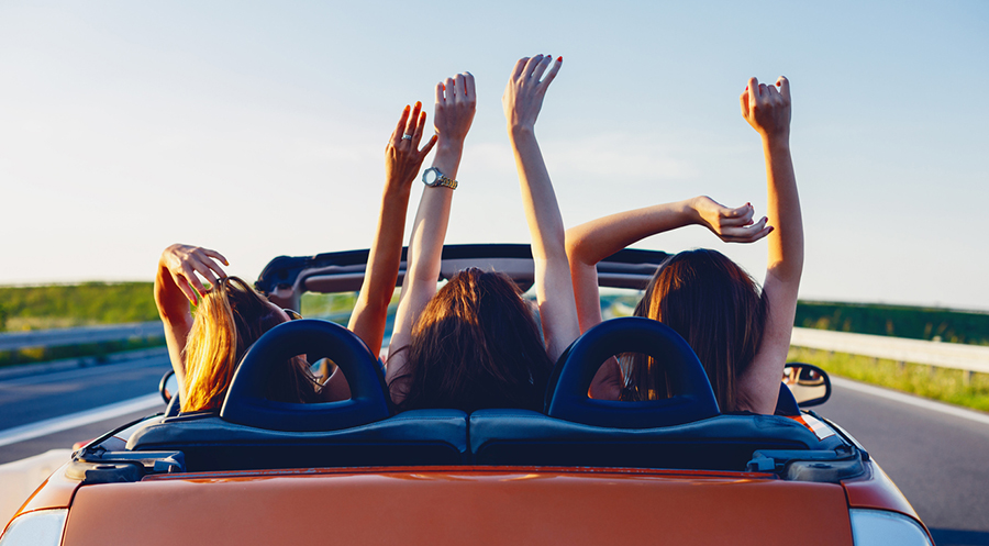Drei junge Frauen fahren im Sommer glücklich in einem orangenen Cabrio mit offenem Dach und strecken dabei die Arme in die Luft.