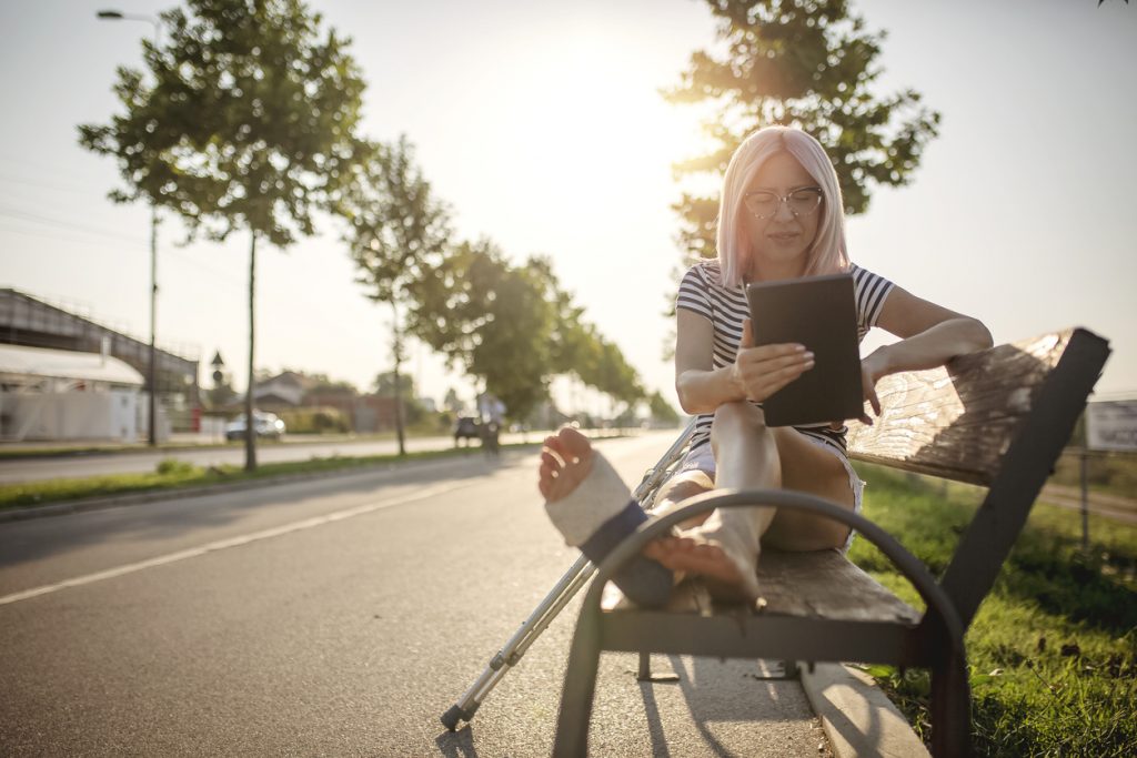 Junge Frau mit Gips am Bein sitzt auf Parkbank und liest