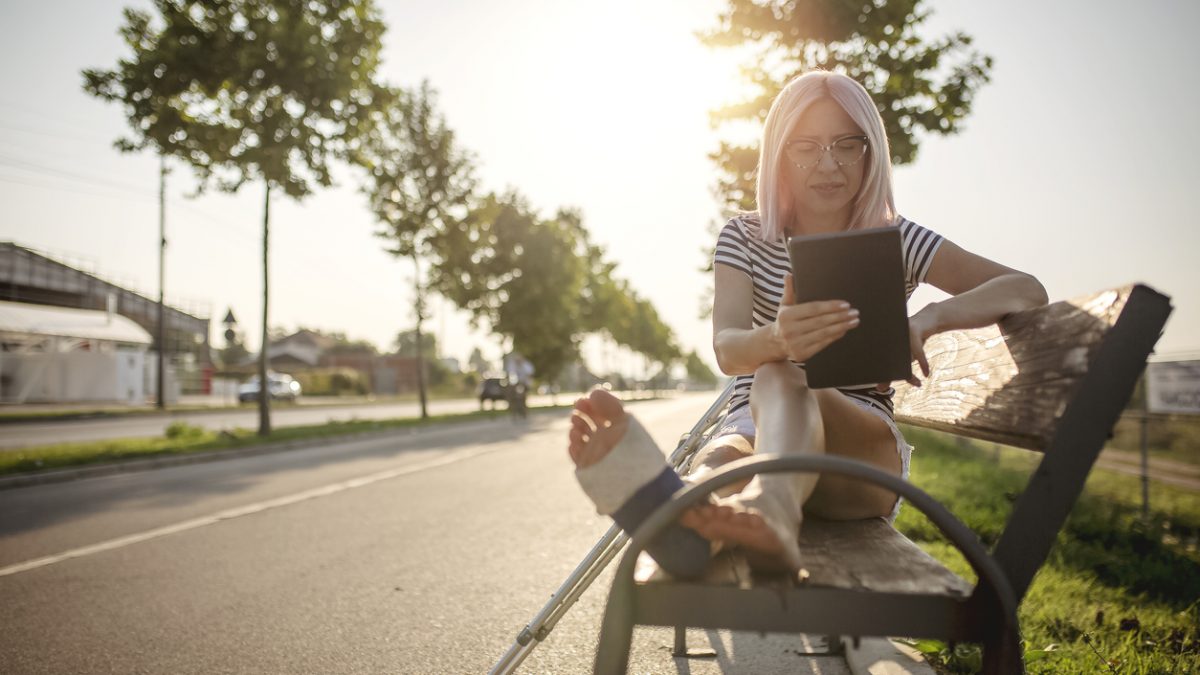 Junge Frau mit Gips am Bein sitzt auf Parkbank und liest