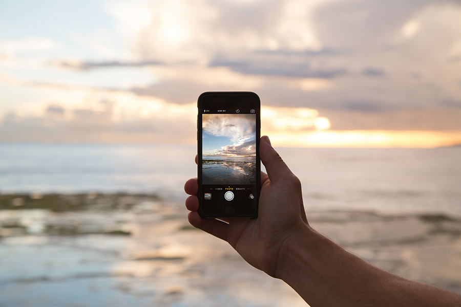 Hand mit Smartphone vor einem Sonnenuntergang hinter dem Meer